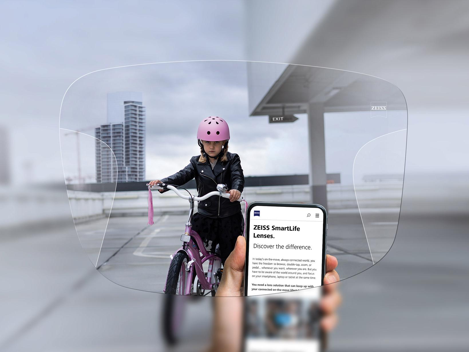 자이스 스마트라이프 누진 렌즈를 통해 휴대폰을 보는 사람. 배경에는 분홍색 보호 헬멧을 쓰고 자전거를 타는 소녀가 보인다. 