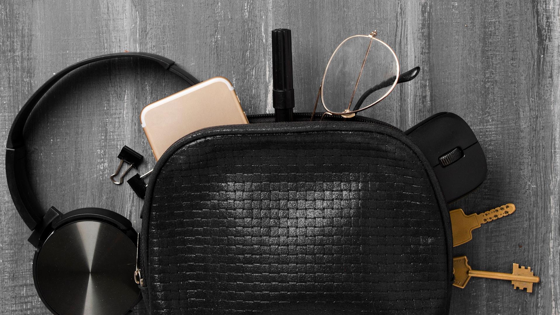 회색 마루에 비스듬히 놓여 있는 검은색 작은 가방에서 헤드폰, 휴대폰, 열쇠, 페이퍼 클립, PC 마우스가 밀려 나와 있고, 자이스 렌즈가 장착되고 듀라비전 코팅이 적용된 안경도 삐져 나와 있다.
