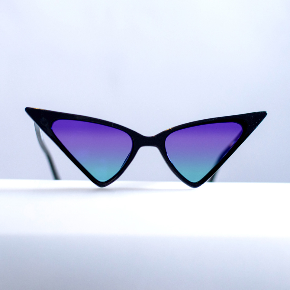 흰색 표면에 기이한 모양의 캣아이 선글라스가 놓여 있다. 청록색에서 보라색으로 변하는 그라데이션 색상이 적용되어 있다