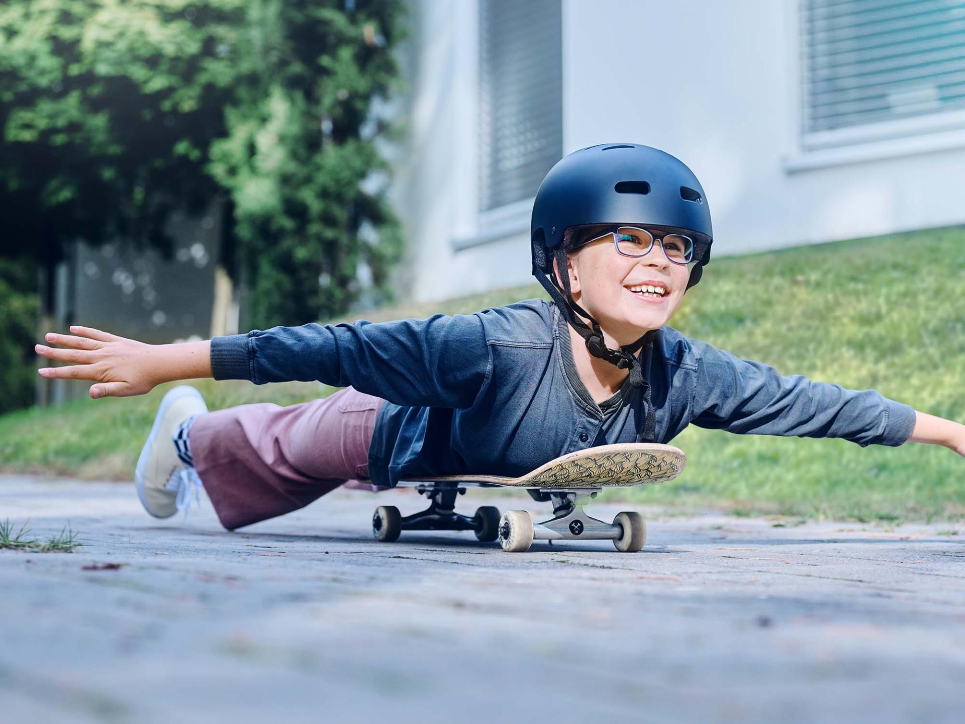 헬멧과 안경을 착용한 소녀가 스케이트 보드에 몸을 얹고 도로를 달리며 두 팔을 활짝 펼치고 있다.