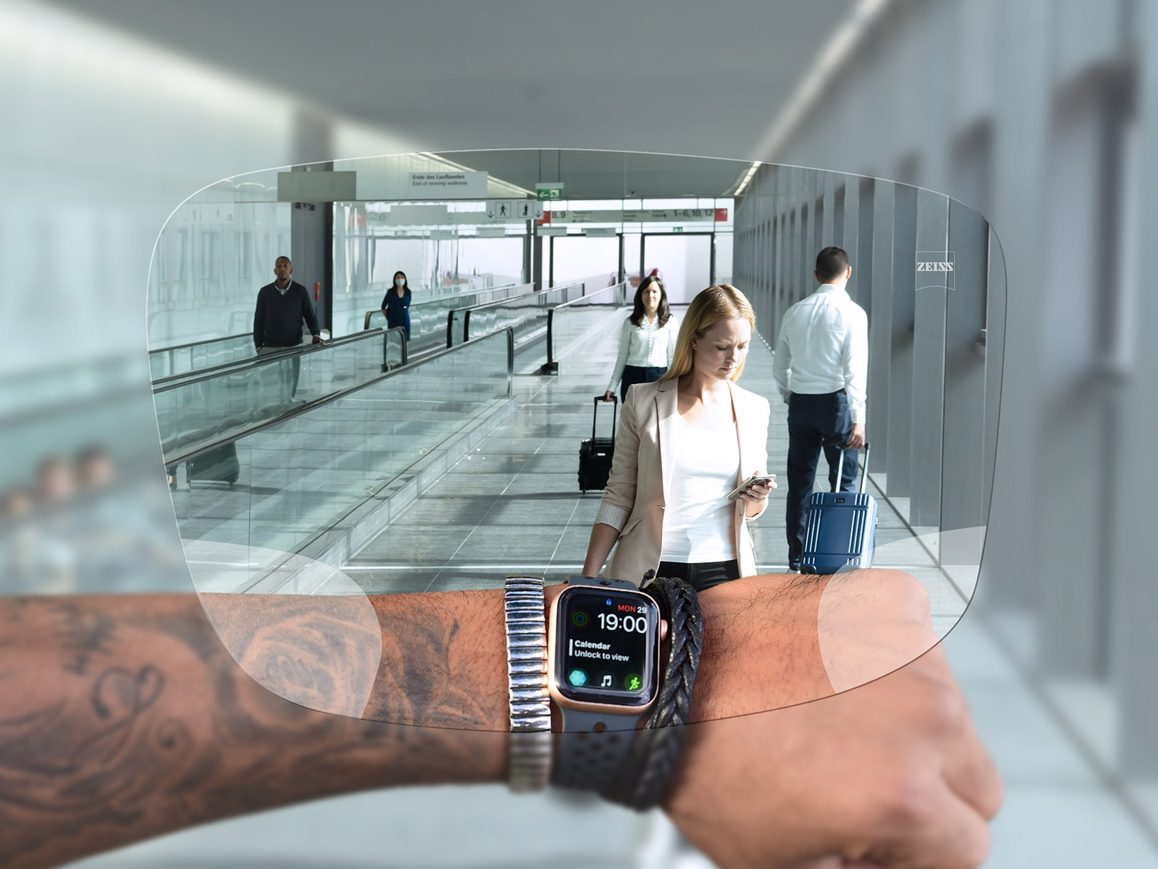 자이스 스마트라이프 디지털 렌즈를 통해 시계를 보는 사람. 배경에는 사람들이 에스컬레이터를 타고 여행 가방을 밀고 있는 전형적인 공항 상황이 보인다. 